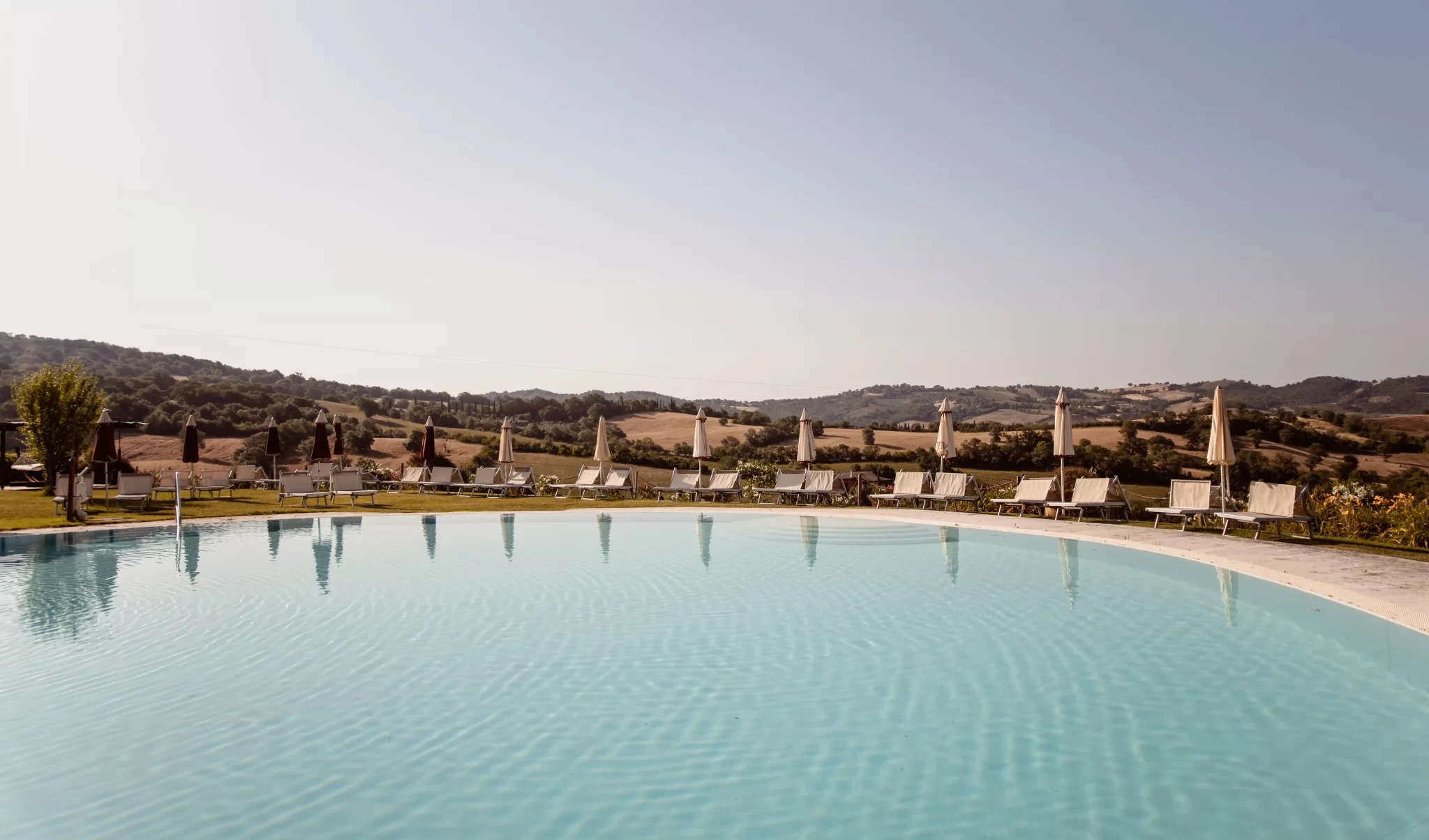 Piscina all'aperto con sdraio e ombrelloni in un paesaggio collinare.