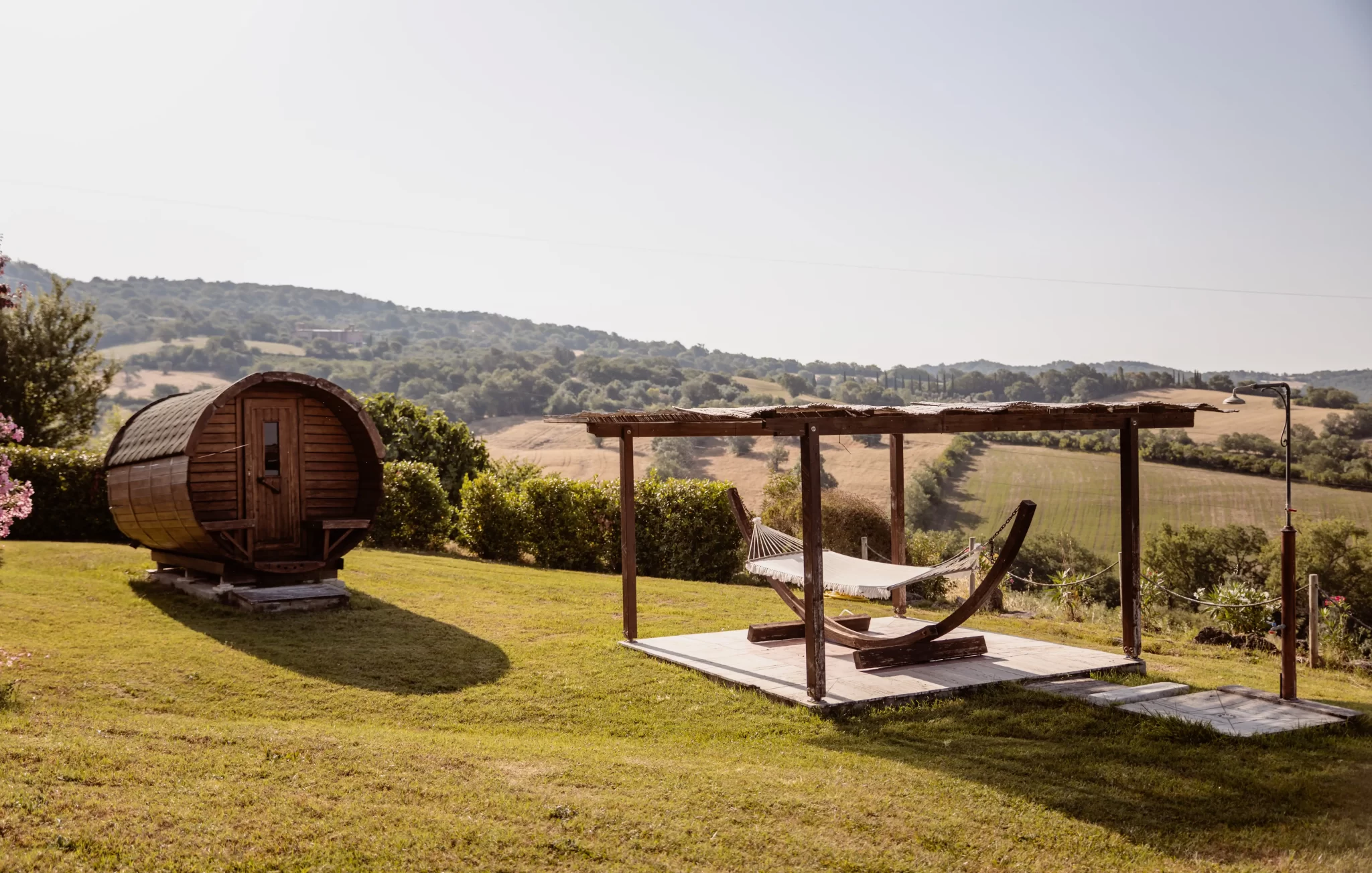 Paesaggio collinare con sauna a botte e amaca sotto un pergolato in legno.