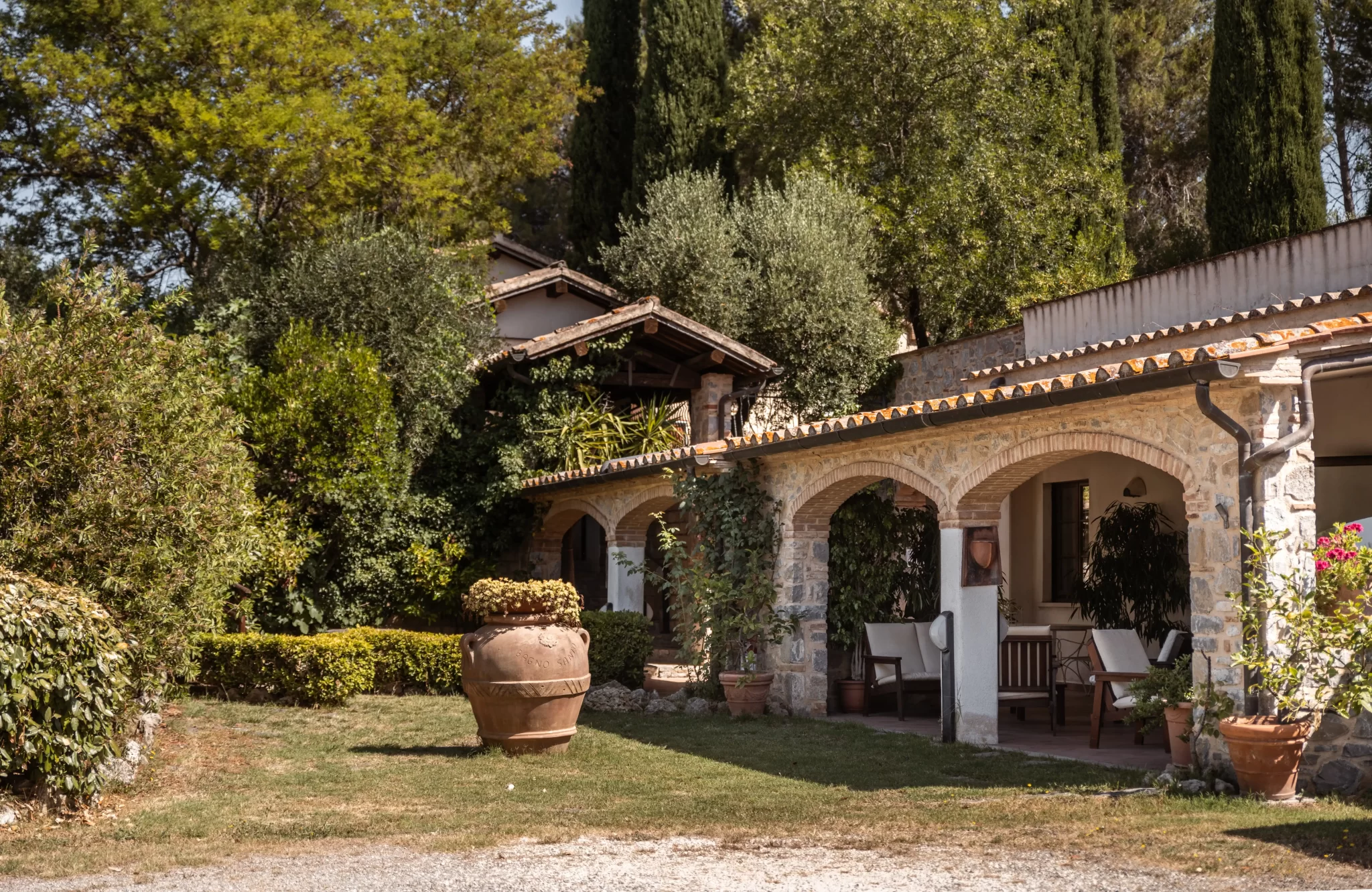 Casale rustico con portico ad arcate, circondato da vegetazione e alberi.
