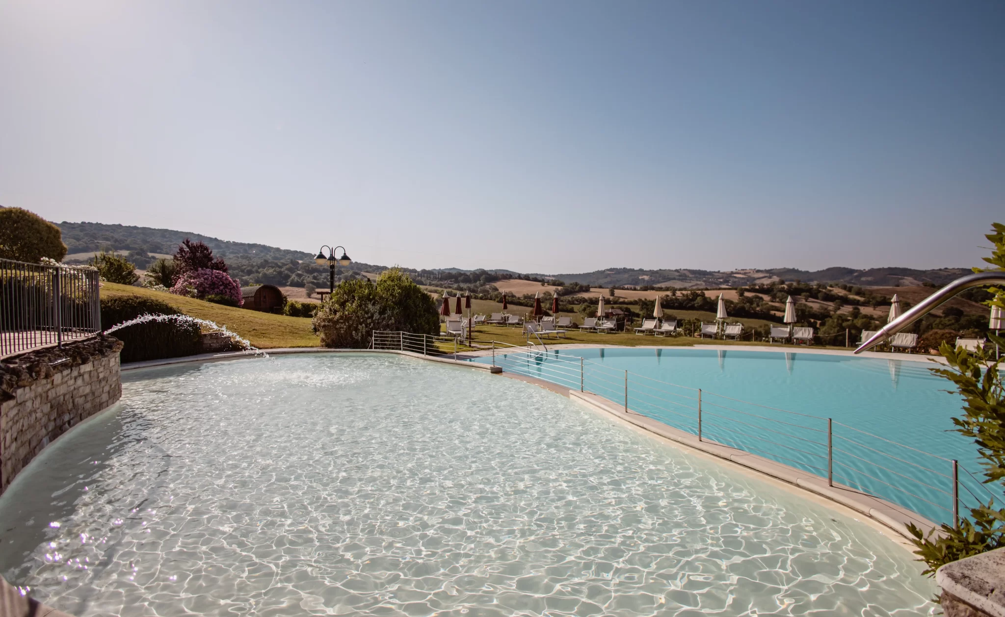 Piscina panoramica immersa nella campagna collinare con sedie a sdraio e ombrelloni.