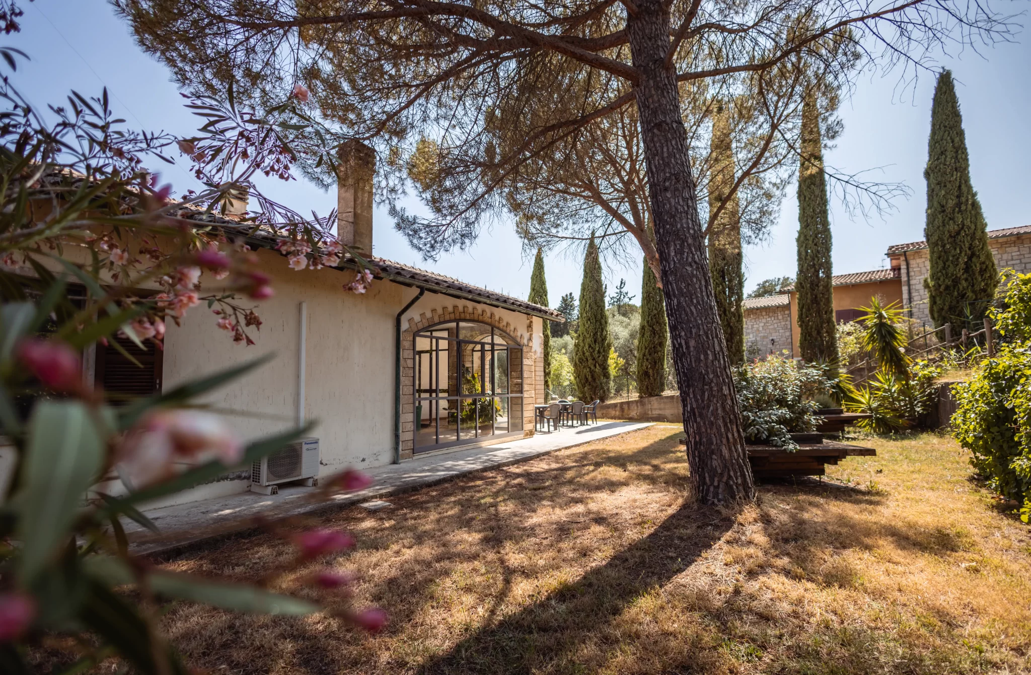 Casa rustica immersa in un giardino, circondata da alberi e piante nel sole.