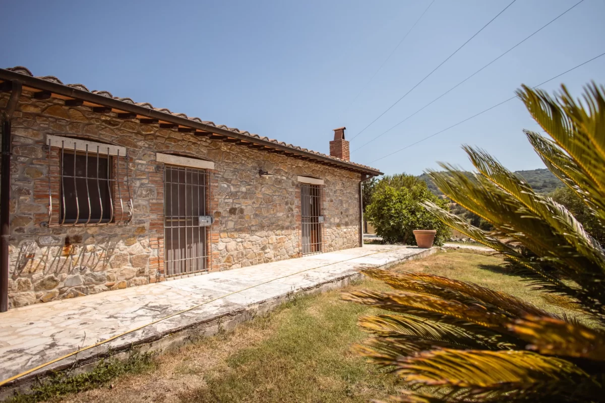 Casa rurale in pietra con tetto in tegole e giardino, in un paesaggio soleggiato.