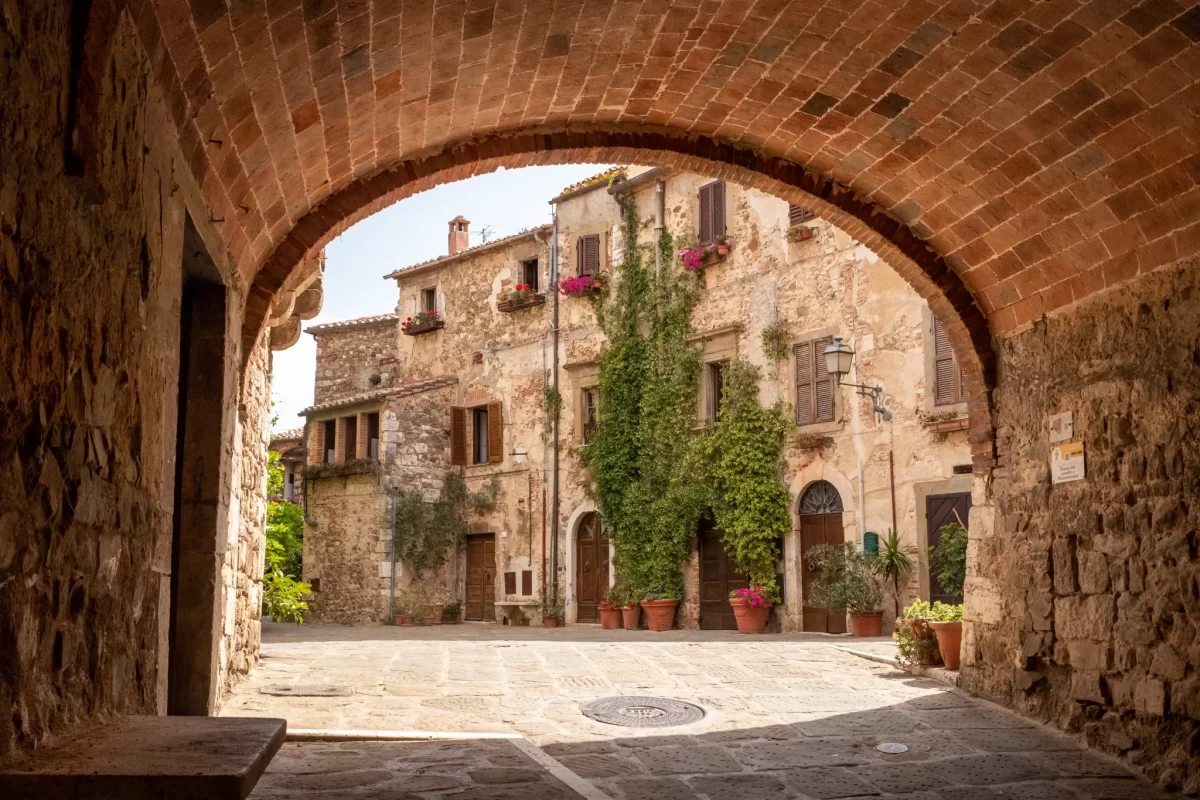 Antico borgo italiano con facciata in pietra e piante rampicanti visto da sotto un arco.