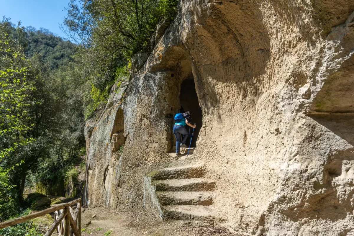 Escursionista entra in una grotta scavata nella roccia lungo un sentiero boschivo.