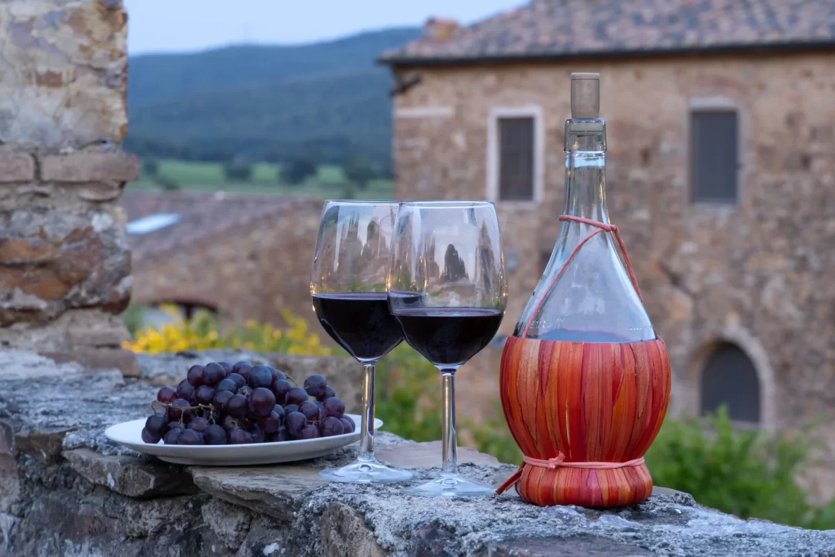Calici di vino rosso e fiasco su un muretto di fronte a un paesaggio rurale italiano.