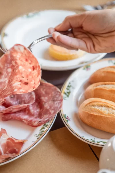 Una colazione con salumi e panini su piatti decorati, con una mano che solleva una fetta di salame, accanto a una tazza di caffè.