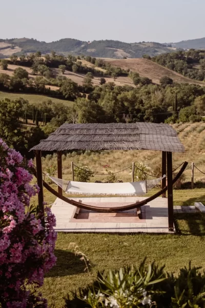 Amaca sotto un pergolato con vista su colline verdi e fiori rosa.