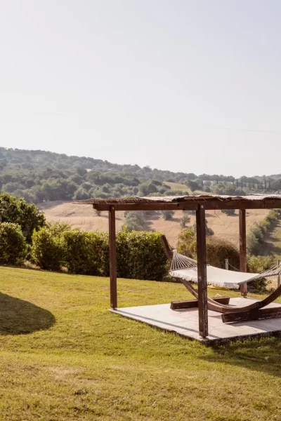 Paesaggio collinare con sauna a botte e amaca sotto un pergolato in legno.