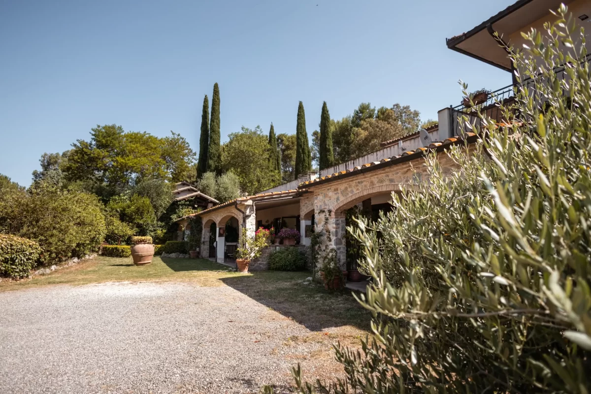 Villa rustica circondata da cipressi e vegetazione rigogliosa sotto un cielo azzurro.