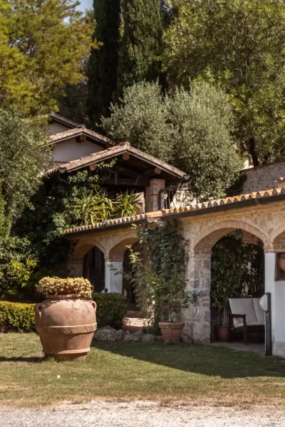 Casale rustico con portico ad arcate, circondato da vegetazione e alberi.