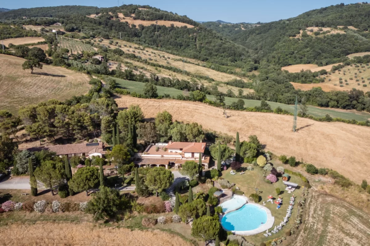 Veduta aerea di una villa con piscina immersa nel paesaggio collinare e verde della campagna.