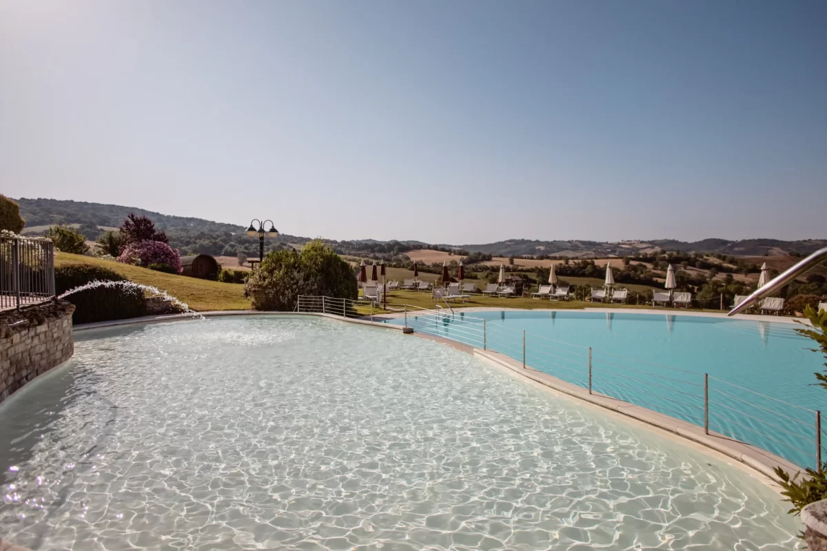 Piscina panoramica immersa nella campagna collinare con sedie a sdraio e ombrelloni.