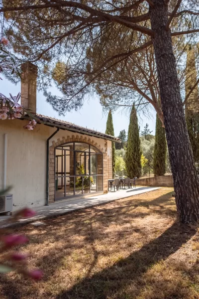 Casa rustica immersa in un giardino, circondata da alberi e piante nel sole.