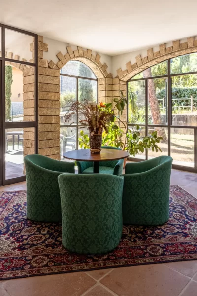 Sala luminosa con ampie vetrate, tavolo rotondo e poltrone verdi su tappeto decorato.