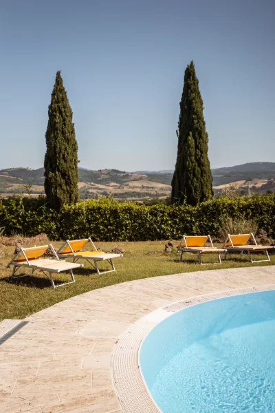 Piscina con lettini prendisole e cipressi sullo sfondo di un paesaggio collinare.