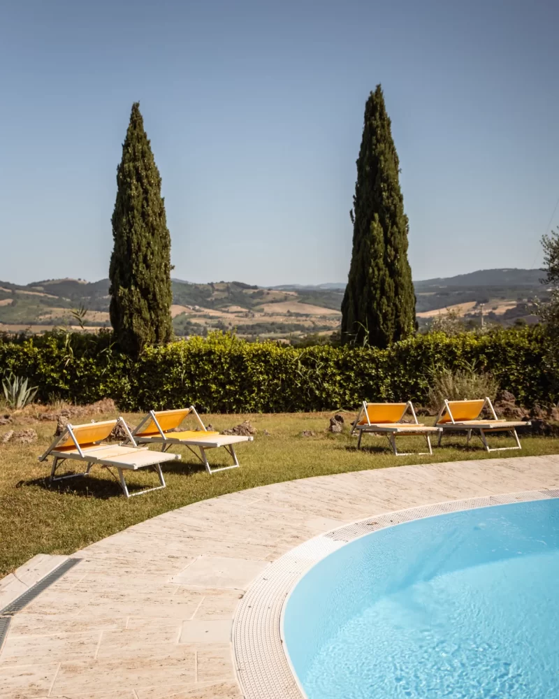 Piscina con lettini prendisole e cipressi sullo sfondo di un paesaggio collinare.