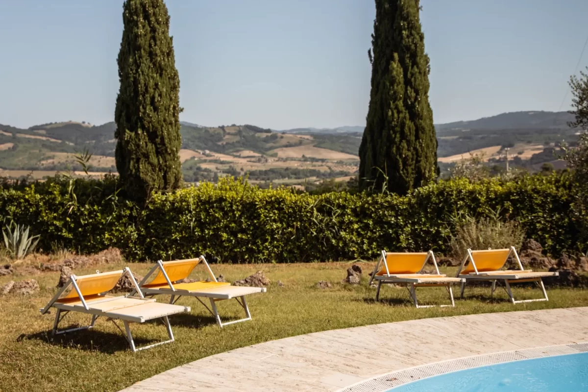 Piscina con lettini prendisole e cipressi sullo sfondo di un paesaggio collinare.